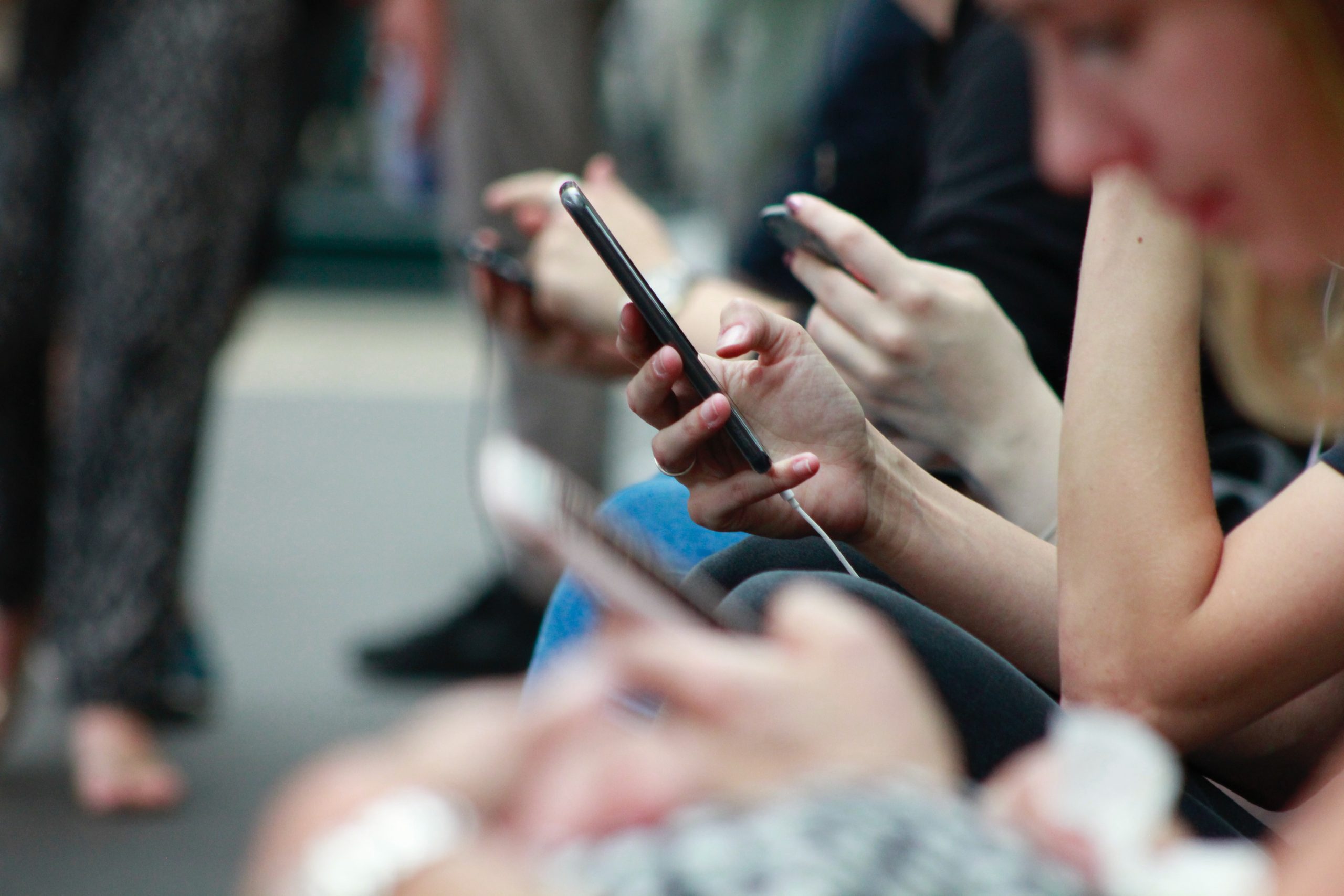 An image of a row of 4 people using their mobiles. The focus of the shot focuses on person holding there smartphone.